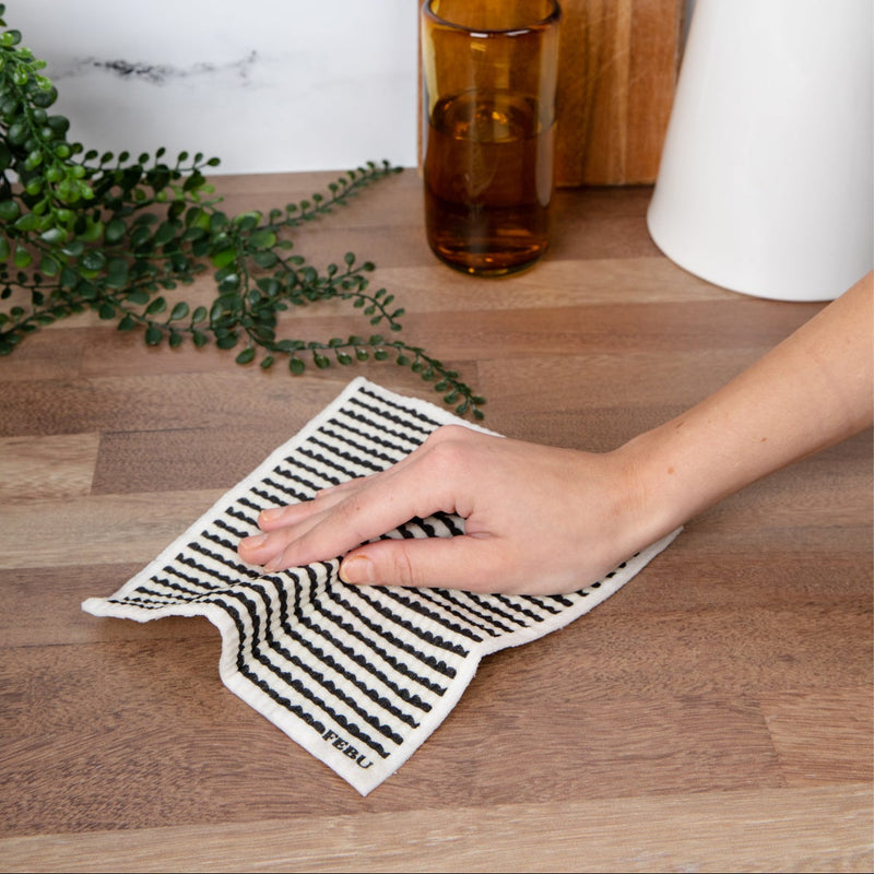 Person using a black and white patterned swedish dish cloth on a wooden surface with kitchen items in the background.